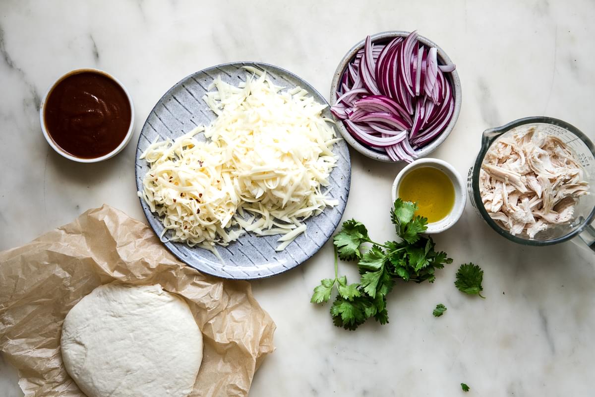 Ingredients for BBQ chicken pizza shown in small bowls including red onions, cilantro, chicken, cheese, bbq sauce and dough.