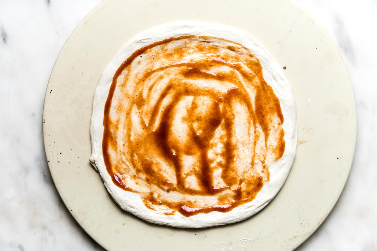 Pizza dough shown on a stone with bbq sauce spread on it.