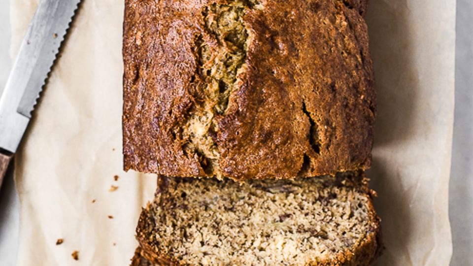 Banana bread loaf sliced on parchment paper shown next to a large knife.