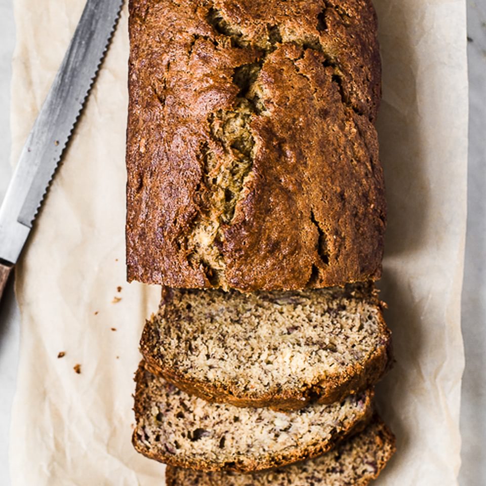 Banana bread loaf sliced on parchment paper shown next to a large knife.