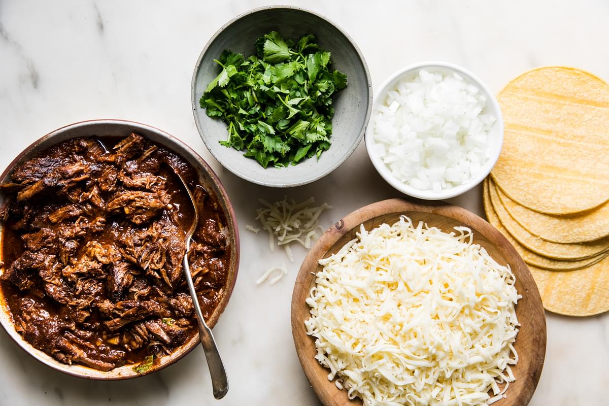 shredded beef birria, cilantro, onions, Oaxaca cheese in bowls on a counter next to a stack of corn tortillas