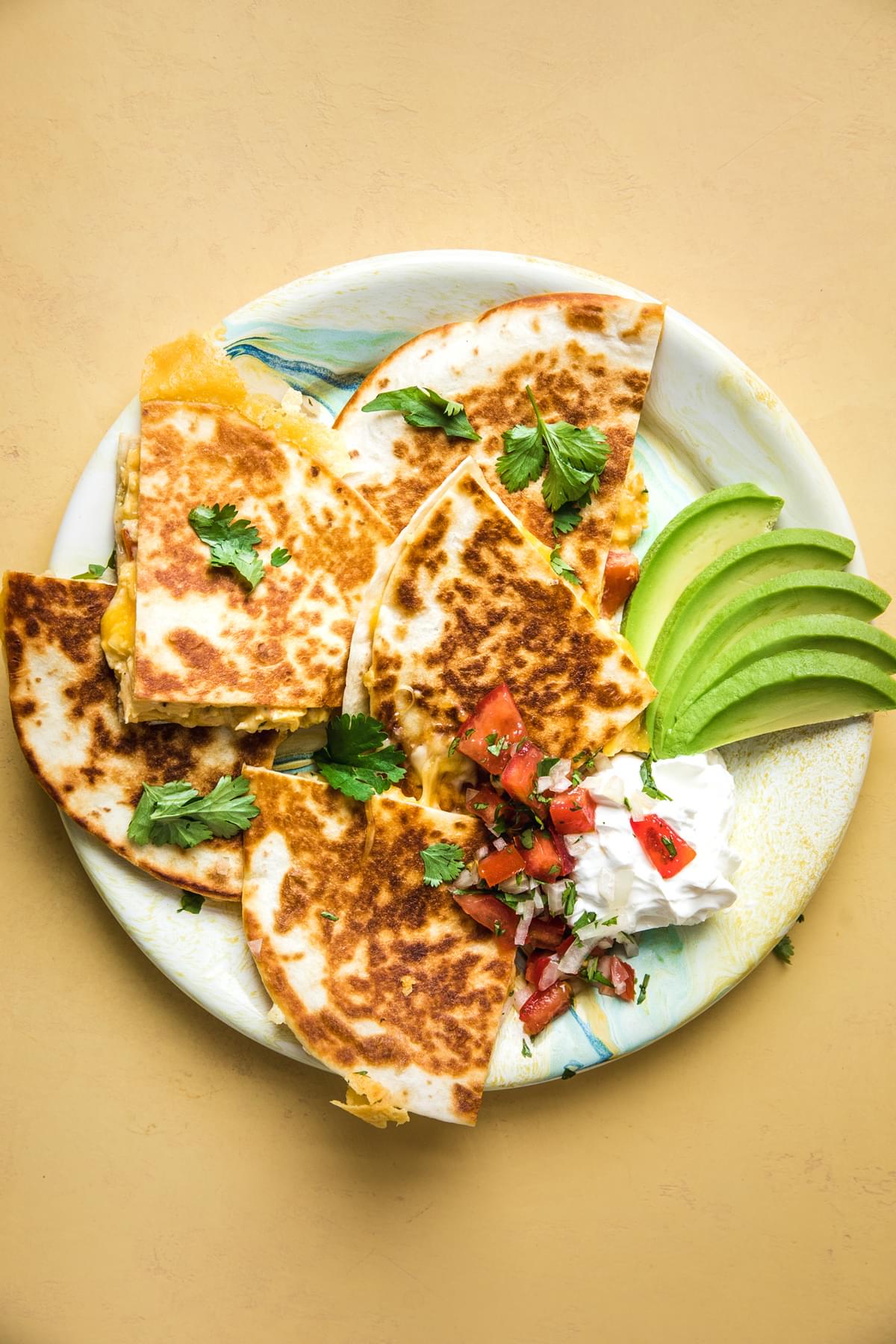 a breakfast quesadilla  filled with scrambled eggs, pico de Gallo, bacon and cheese on a plate with avocado and sour cream