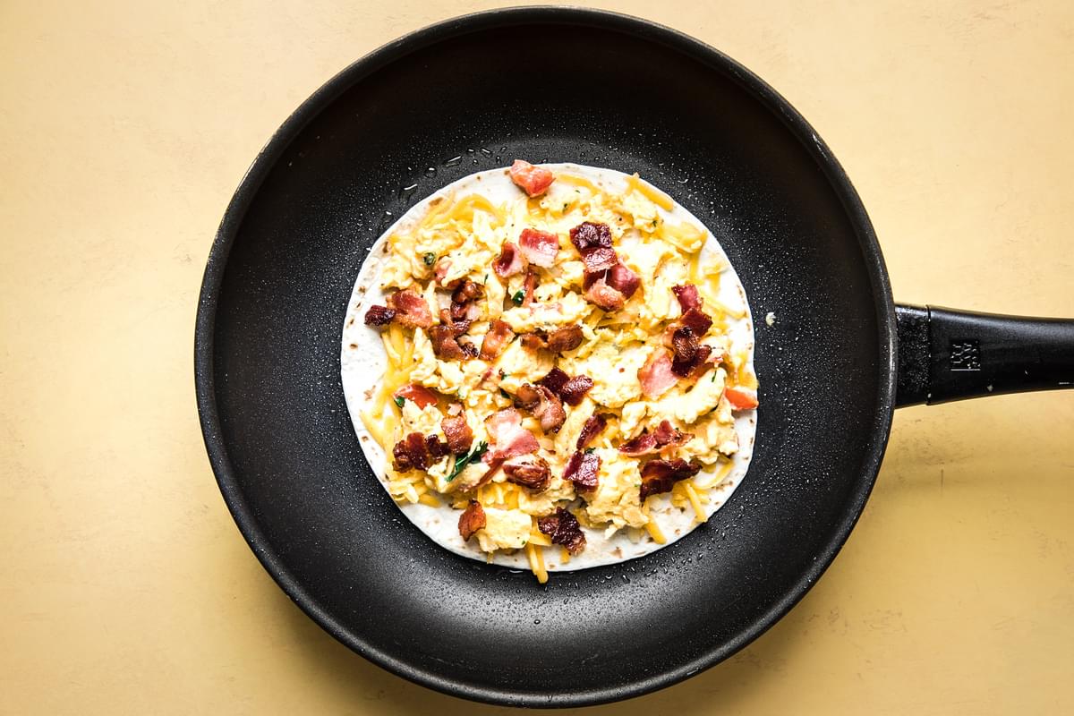 a four tortilla topped with scrambled eggs, pico de Gallo, cheese and bacon in a skillet to make a breakfast quesadilla