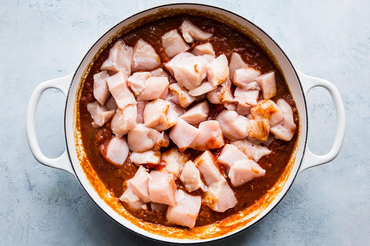 raw chicken being added to butter chicken sauce in a large white pot.