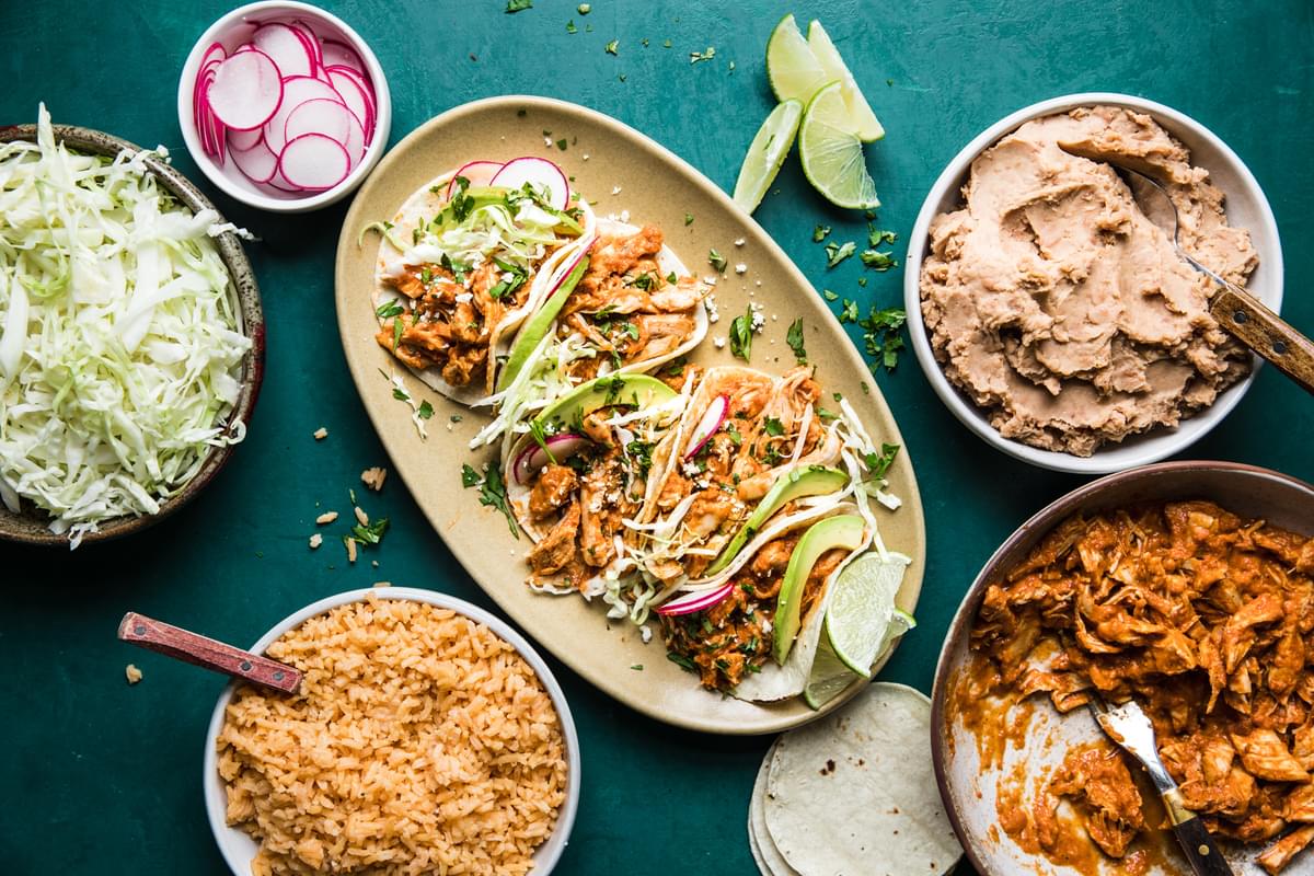 chicken Tinga tacos on a plate next to bowls of beans and rice served with lime, radishes and shredded lettuce