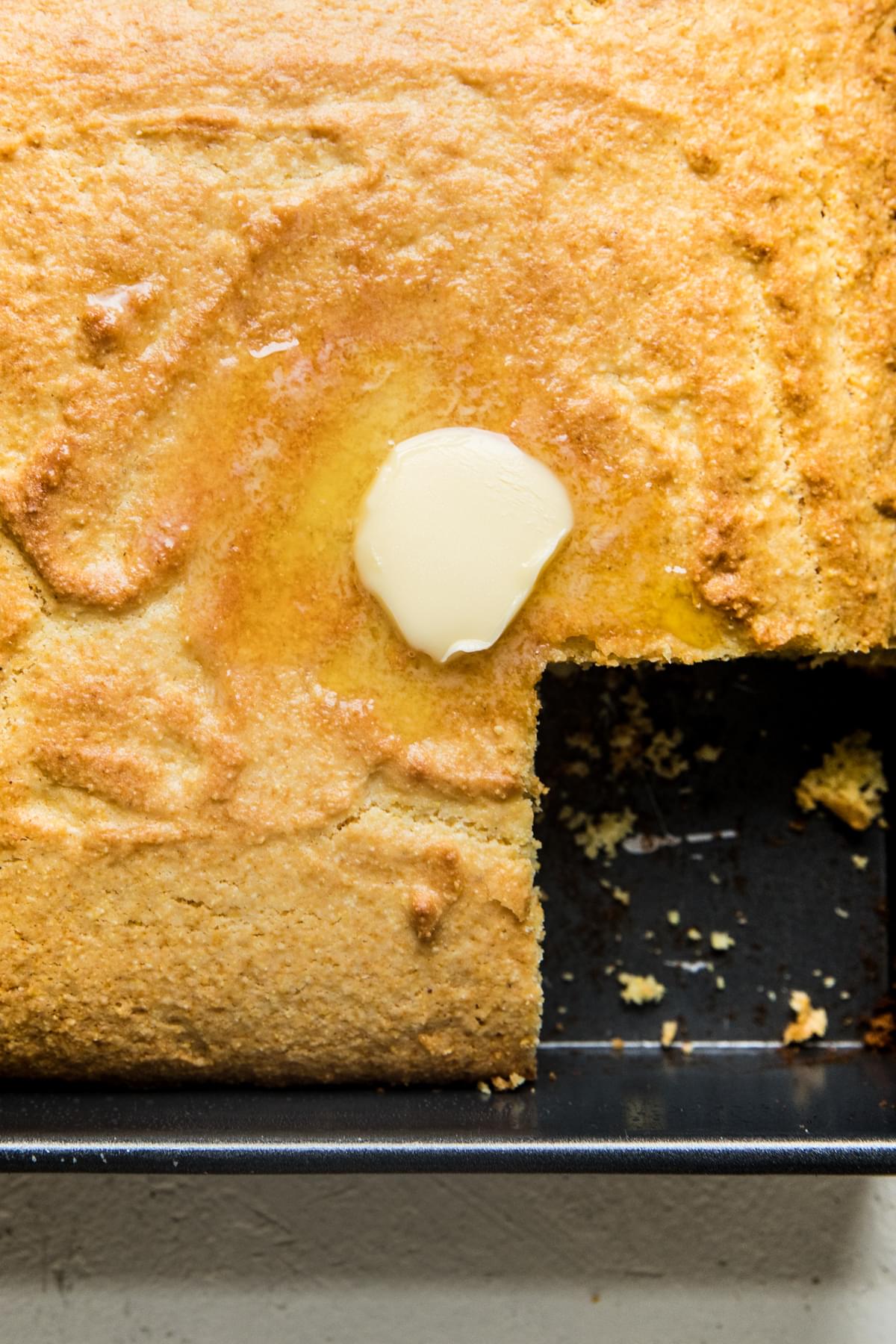 homemade corn bread in a baking dish with a slice taken out and a pat of butter
