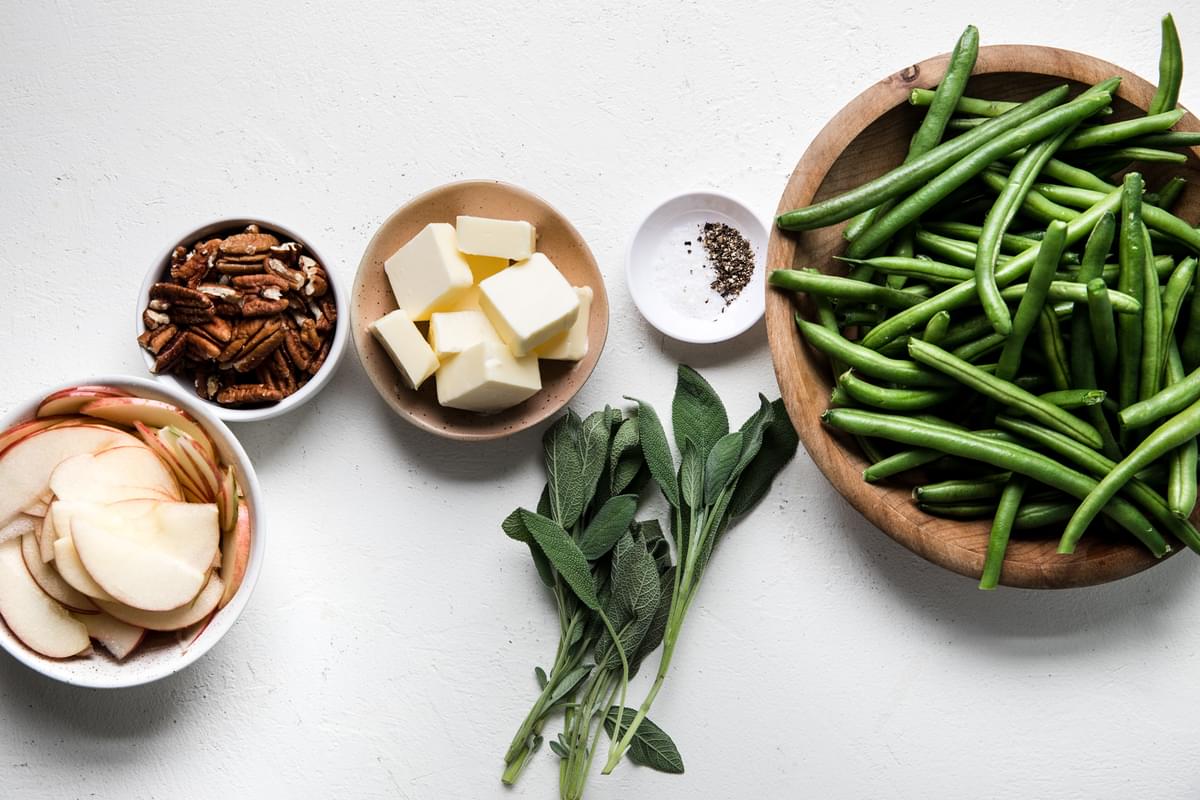 fresh green beans, sliced apples, butter, pecans, salt, pepper and sage leaves in bowls on the counter