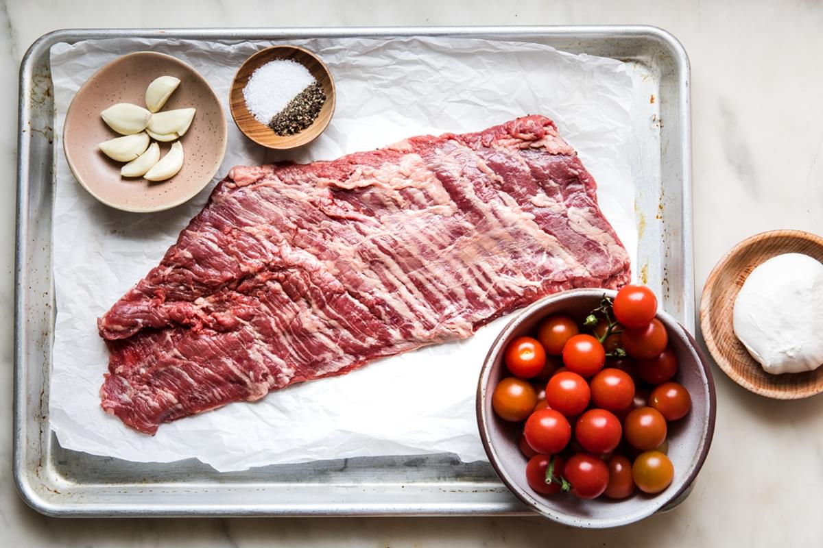 raw skirt steak, a bowl of cherry tomatoes, salt, pepper, burrata cheese and garlic on a sheet pan