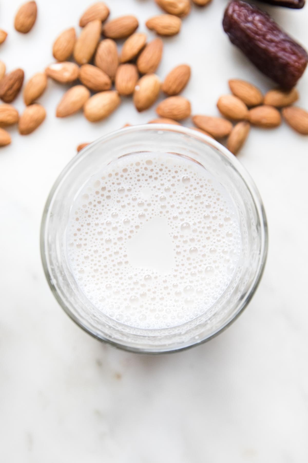 a glass of homemade almond milk sitting on the counter surrounded by almonds and dates