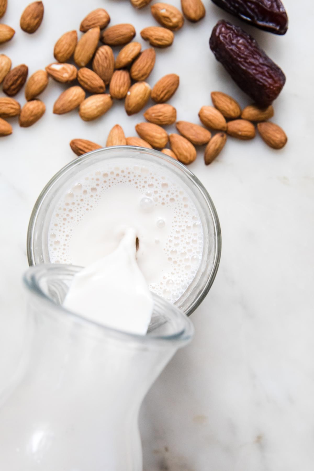 a carafe of homemade almond milk being poured into a glass surrounded by almonds and dates on the counter
