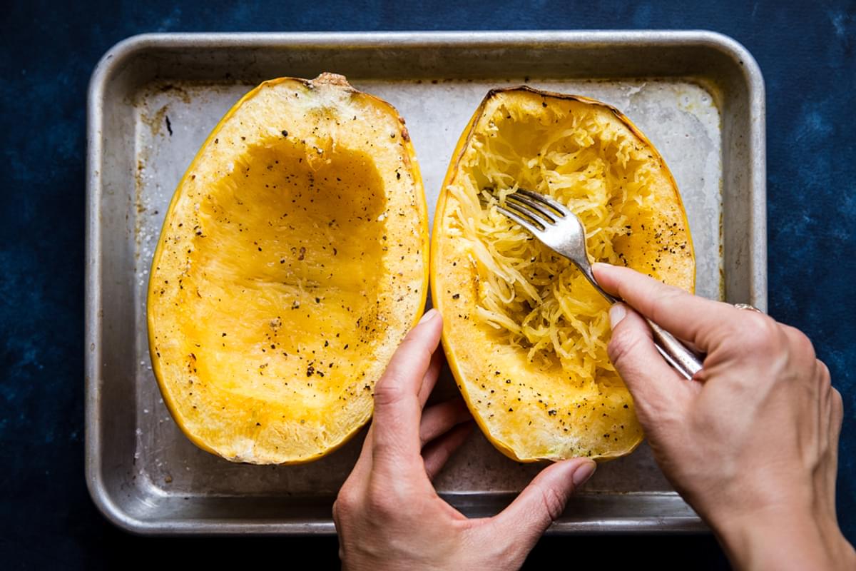 oven roasted spaghetti squash with salt and pepper on a baking sheet with hands using a fork to scrape some out