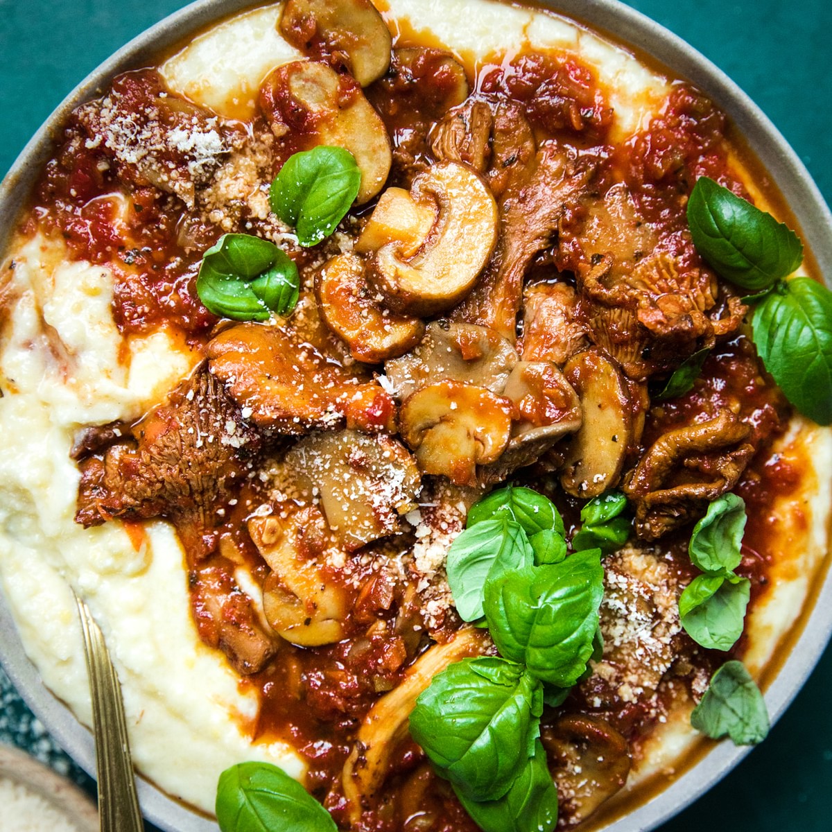 a bowl of mushroom ragù served over creamy polenta topped with basil leaves next to a bowl of grated parmesan