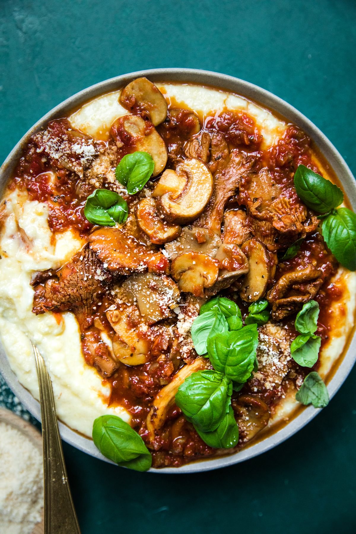 a bowl of mushroom ragù served over creamy polenta topped with basil leaves next to a bowl of grated parmesan
