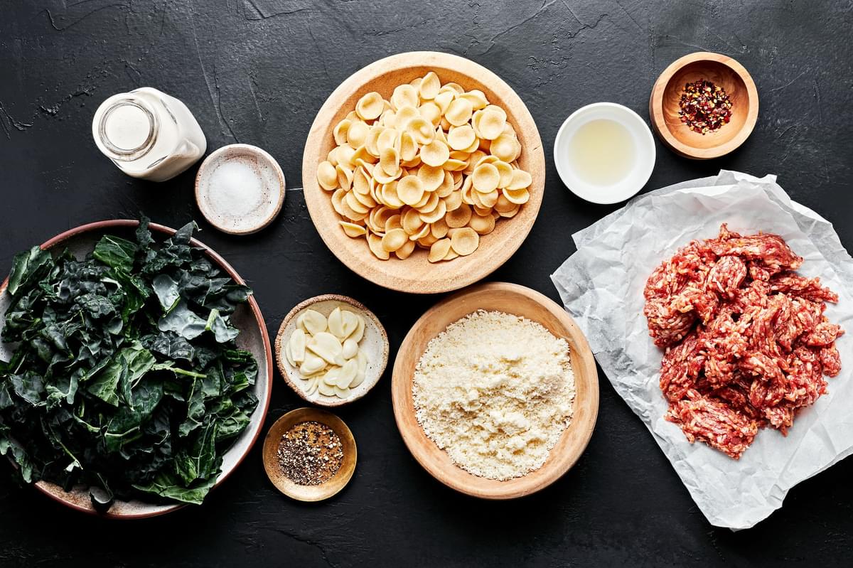 ingredients to make orecchiette pasta in small bowls including kale, sausage, parmesan cheese, sausage, pasta and pepper.