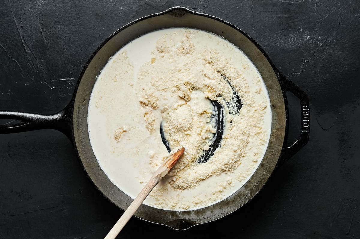 Heavy cream and parmesan being stirred with a wooden spoon in a black cast iron skillet