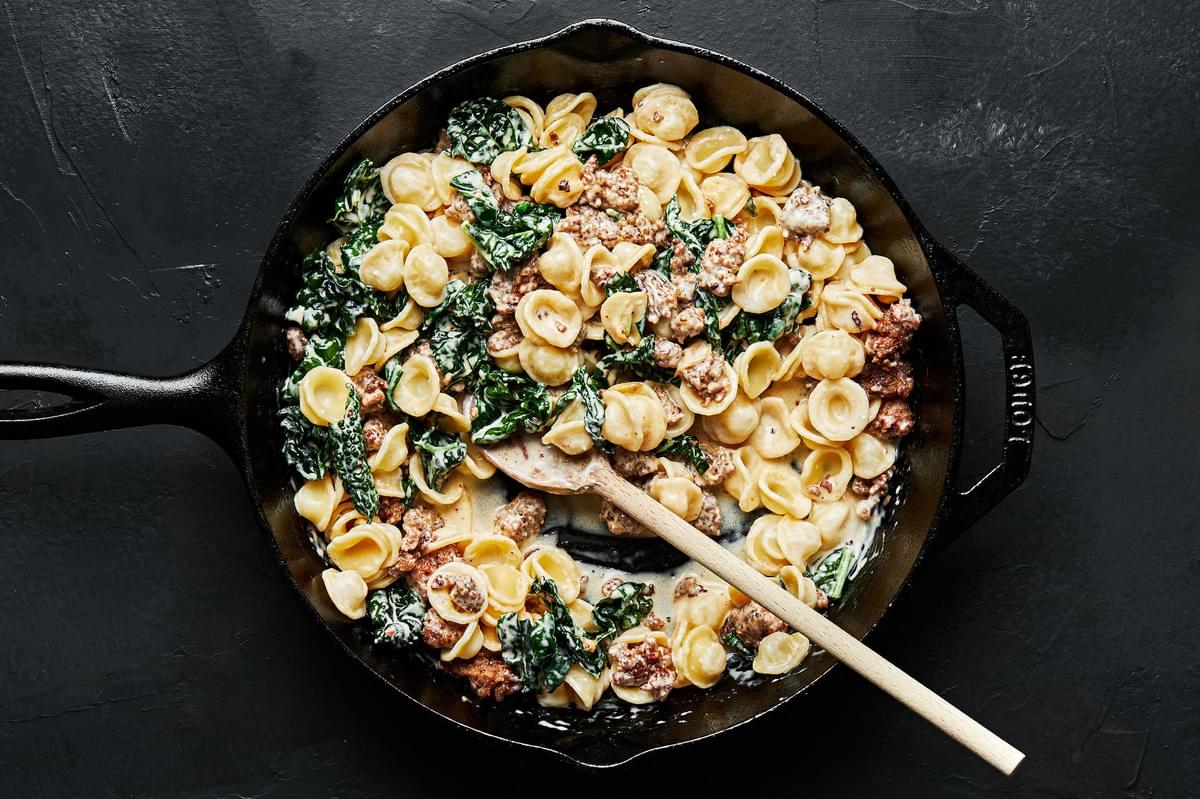 orecchiette pasta with sausage and kale in a large black skillet with a wooden spoon