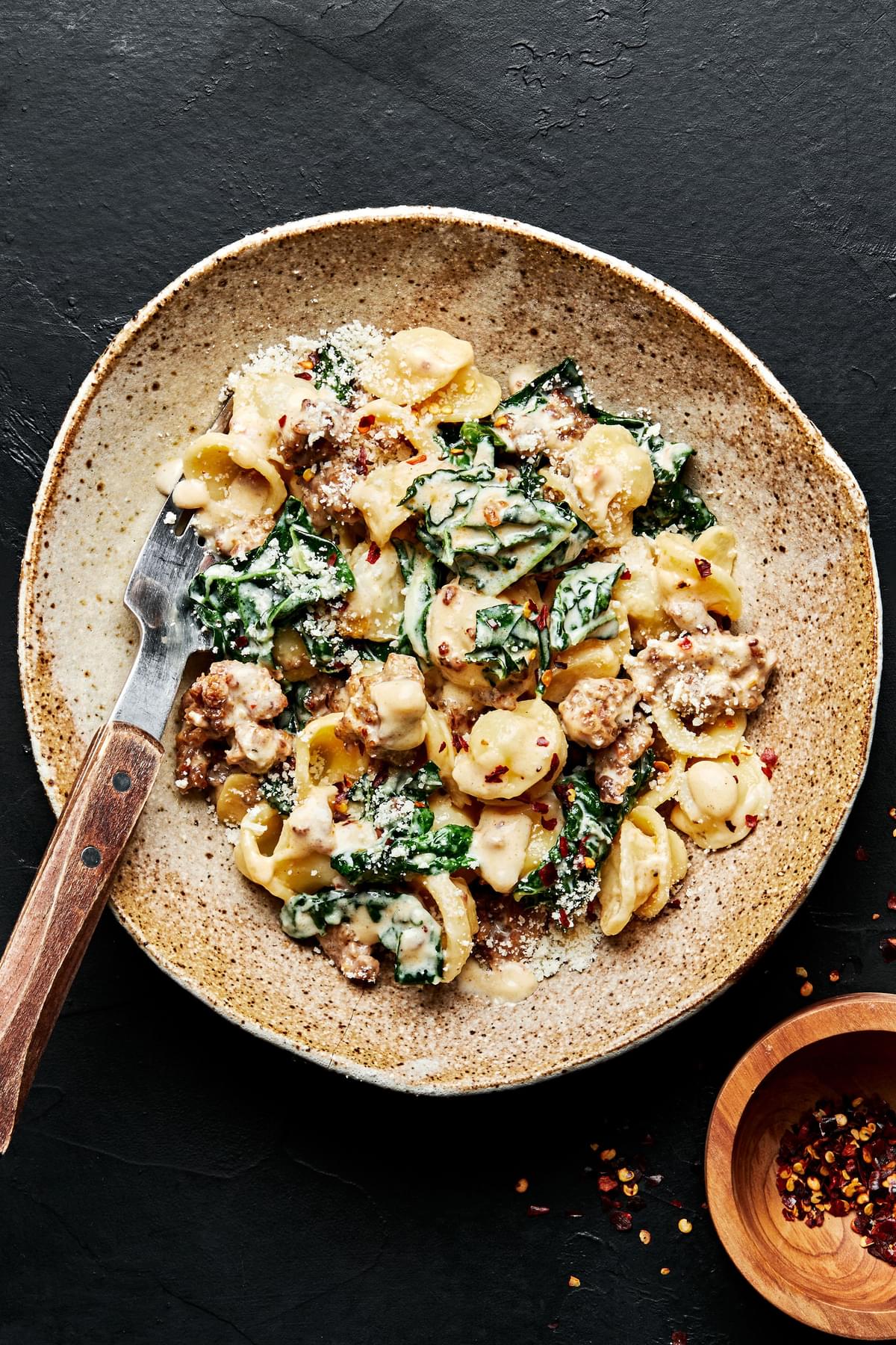 orecchiette pasta with kale and sausage in a bowl next to a small bowl with red pepper flakes