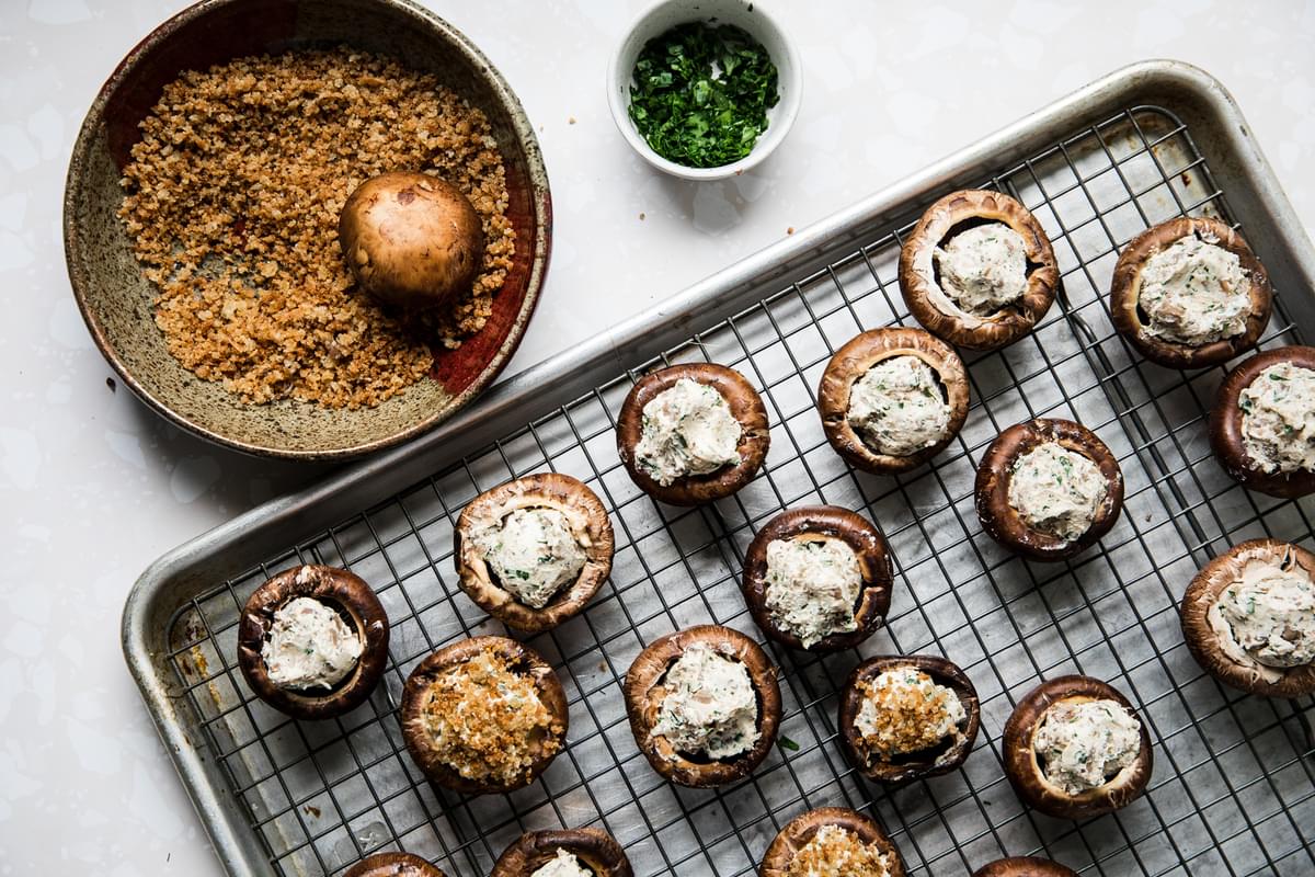 raw stuffed mushrooms on a baking sheet being dipped in a bowl of panko breadcrumbs
