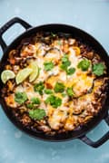 Homemade vegetarian enchilada skillet topped with cheese, cilantro and lime wedges in a skillet on the counter