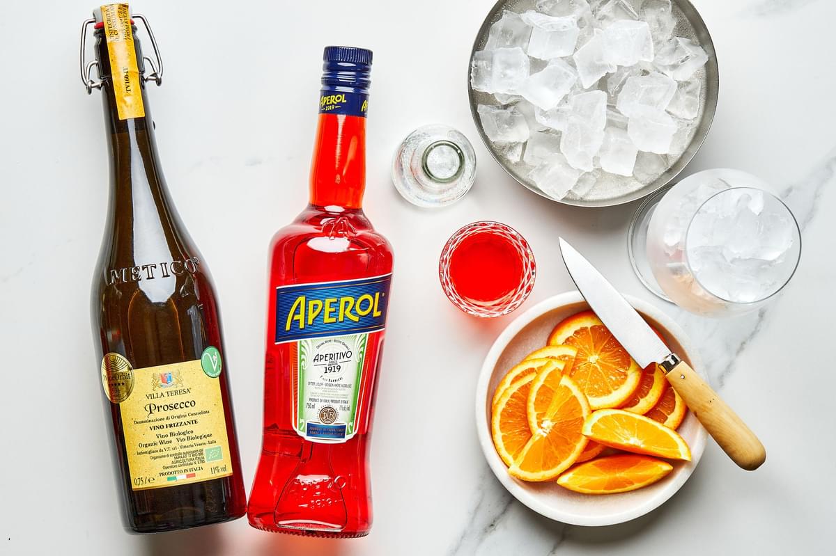 bottles of prosecco and and aperol, a bowl of ice, a stemmed wine glass filled with ice, orange slices and a cocktail jigger