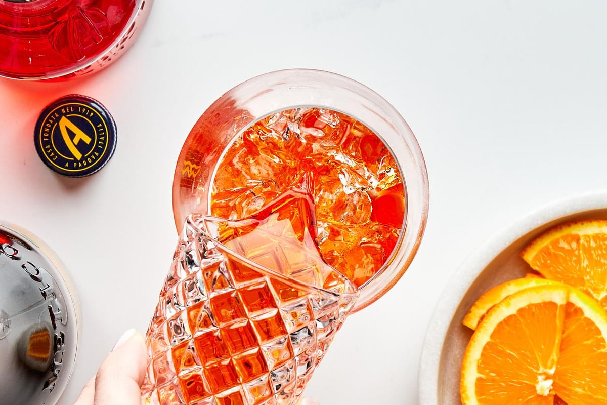 Aperol being poured over ice into a wine glass next to bottles of prosecco and aperol and bowl of orange slices