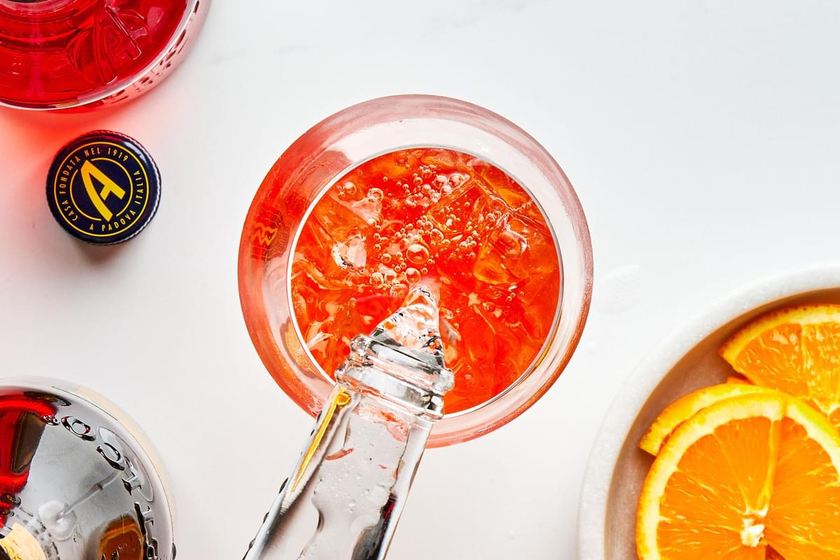 Prosecco being poured over ice and aperol into a wine glass next to bottles of prosecco and aperol and bowl of orange slices