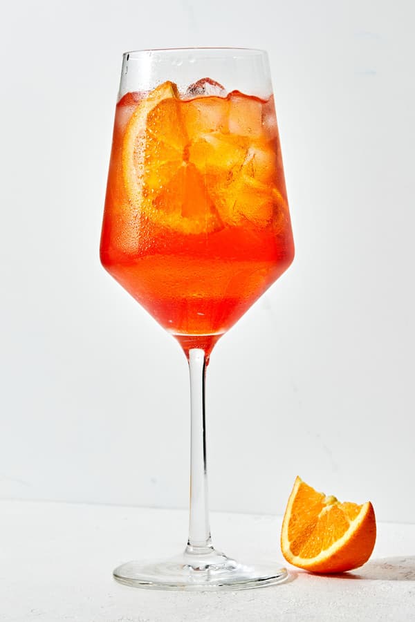 an Aperol Spritz in a stemmed glass over ice with an orange slice as garnish and an orange slice on the counter beside it