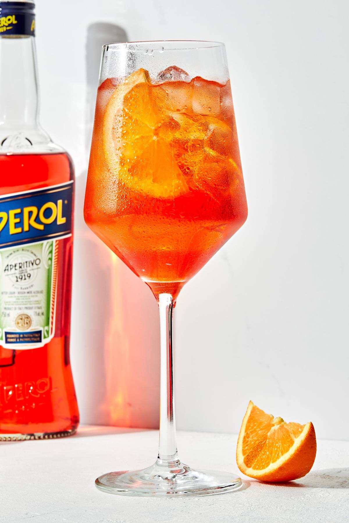 an Aperol Spritz in a stemmed glass over ice with an orange slice as garnish with prosecco and aperol bottles beside it