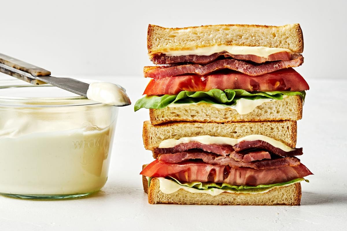 two halves of a BLT with homemade mayonnaise stacked on top of each other next to a glass bowl of homemade mayonnaise
