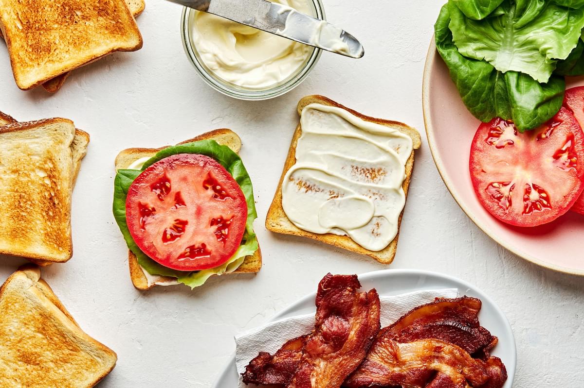 a BLT being assembled surrounded by toast, bacon, homemade mayonnaise, sliced tomato and lettuce