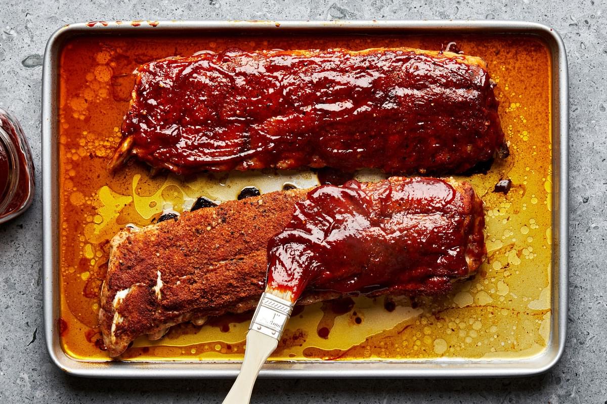 baked pork ribs on a baking sheet being slathered with homemade bbq sauce