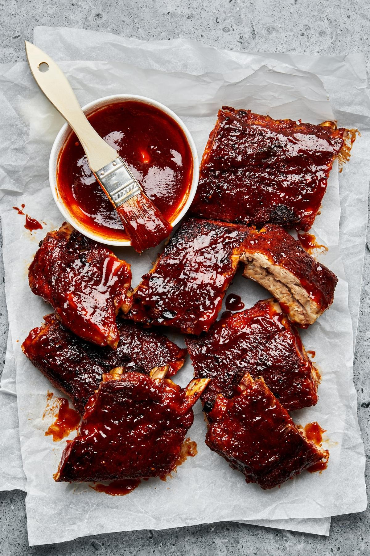 baked pork ribs covered with homemade bbq sauce next to a bowl of extra bbq sauce with a pastry brush in it