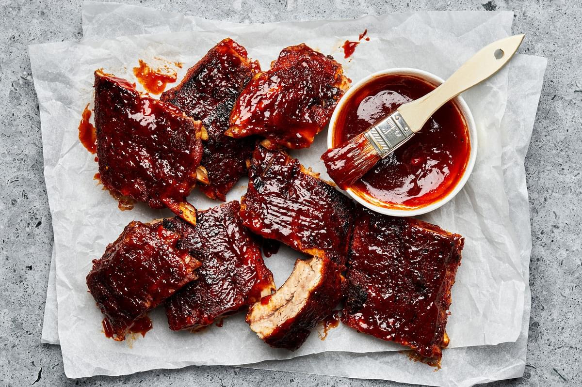 baked pork ribs covered with homemade bbq sauce next to a bowl of extra bbq sauce with a pastry brush in it