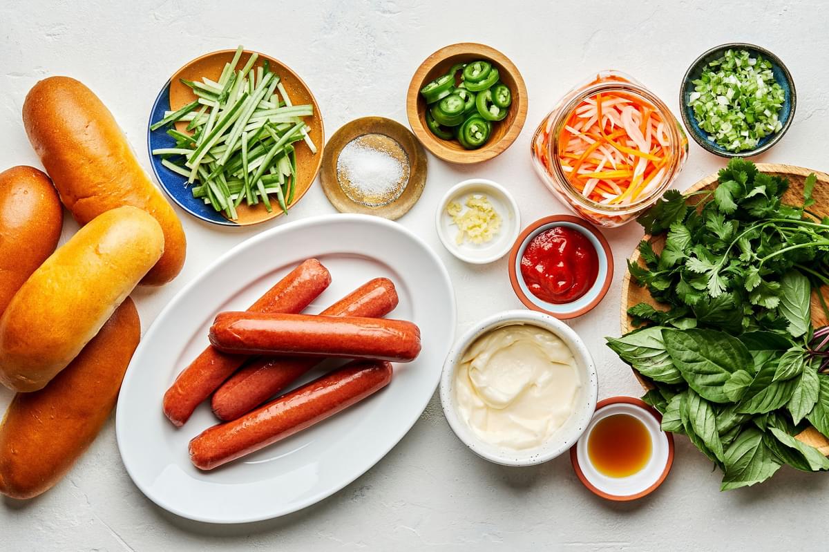 hot dogs, buns,  pickled veggies, cilantro, basil, cucumbers, jalapeños and ingredients for sriracha mayo in bowls