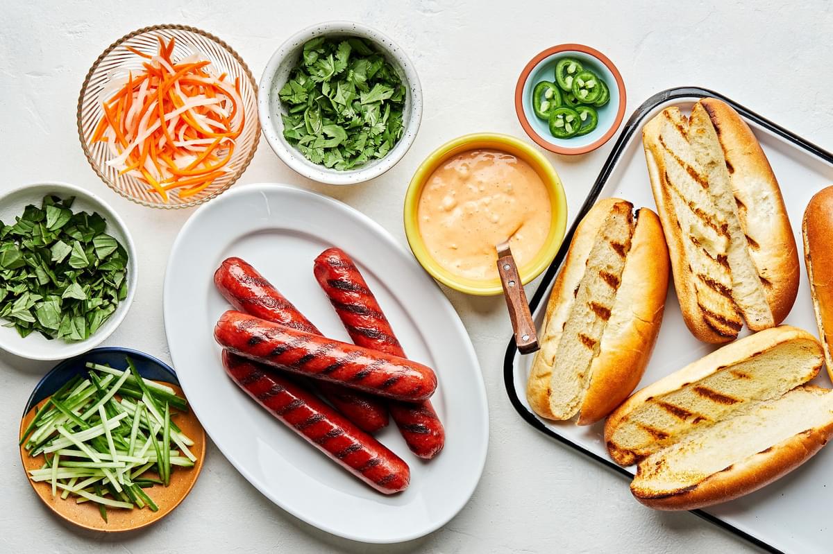 Banh Mi Hot Dogs ingredients in bowls. Sriracha mayo, pickled veggies, cilantro, basil, cucumbers, jalapeños and buns