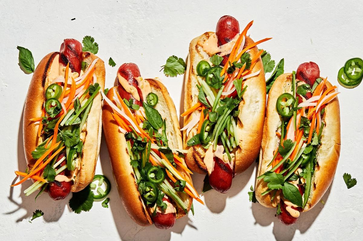 Banh Mi Hot Dogs drizzled with sriracha mayo topped with pickled veggies, cilantro, basil, cucumbers and jalapeños