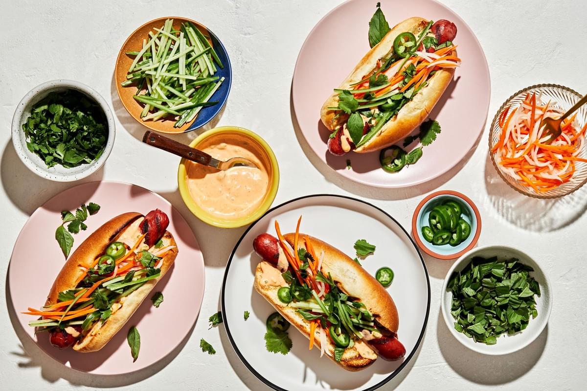 Banh Mi Hot Dogs surrounded by fixings, Sriracha mayo, pickled veggies, cilantro, basil, cucumbers and jalapeños