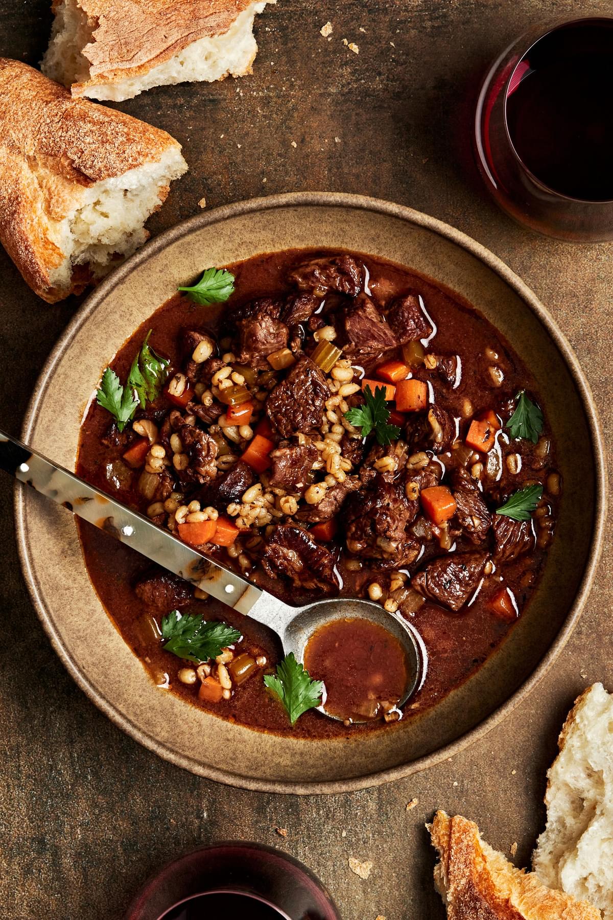 a bowl of homemade beef & barley soup with a spoon surrounded by crusty bread for serving