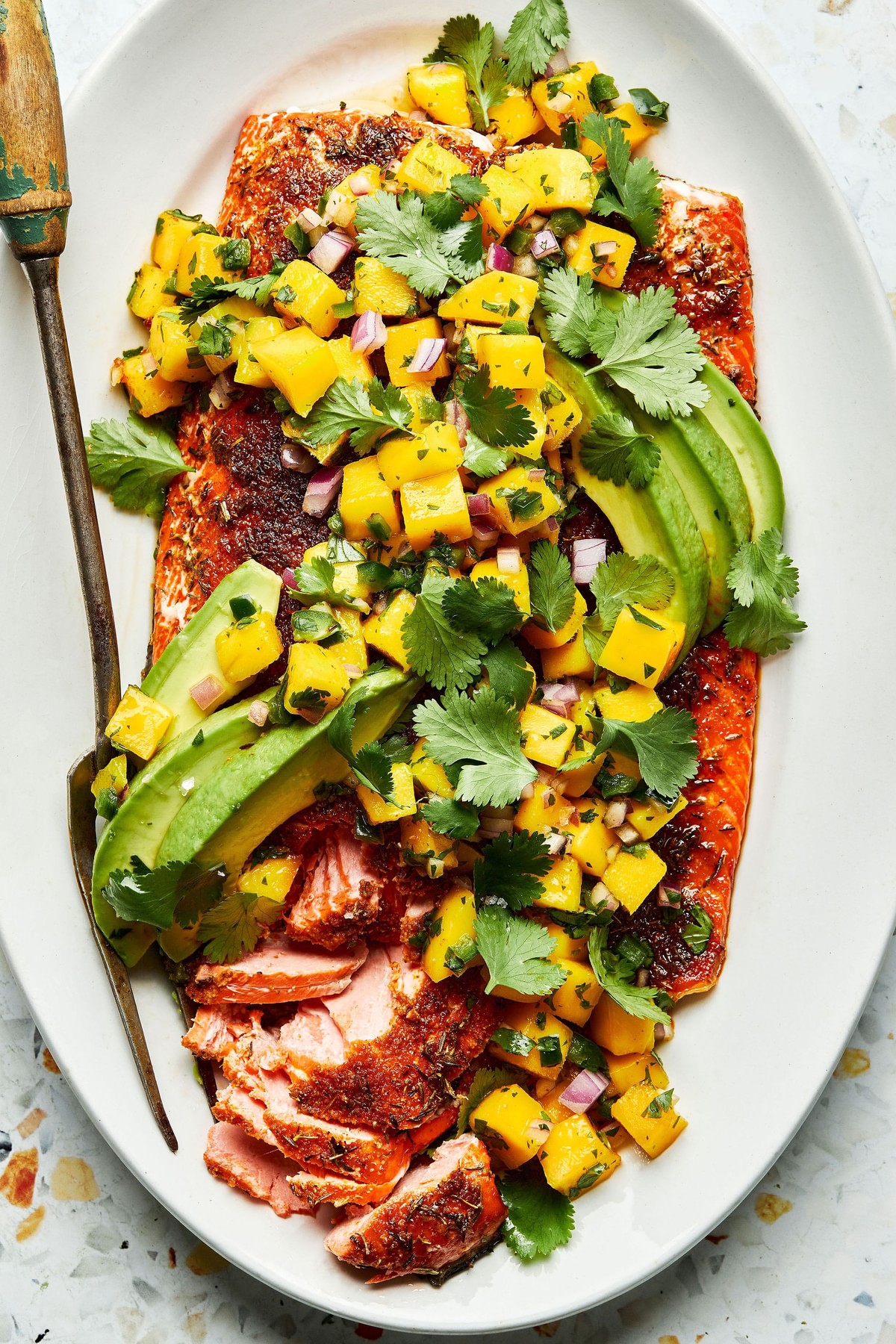 blackened salmon topped with homemade mango salsa and avocado on a serving plate with a serving fork