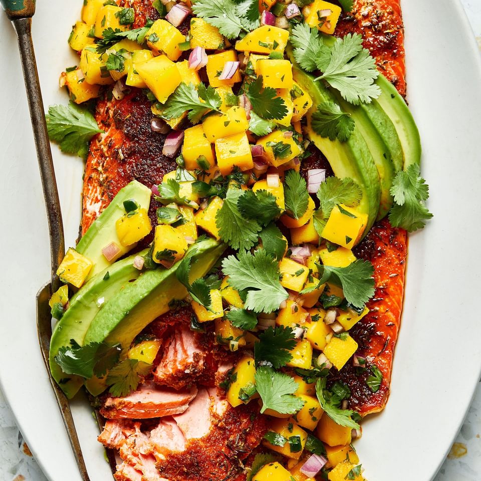 blackened salmon topped with homemade mango salsa and avocado on a serving plate with a serving fork