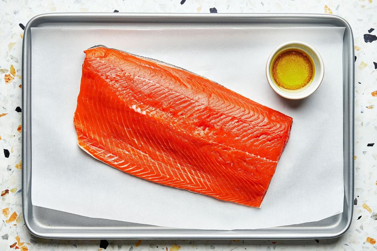 a salmon filet on a parchment paper lined baking sheet next to a bowl of olive oil