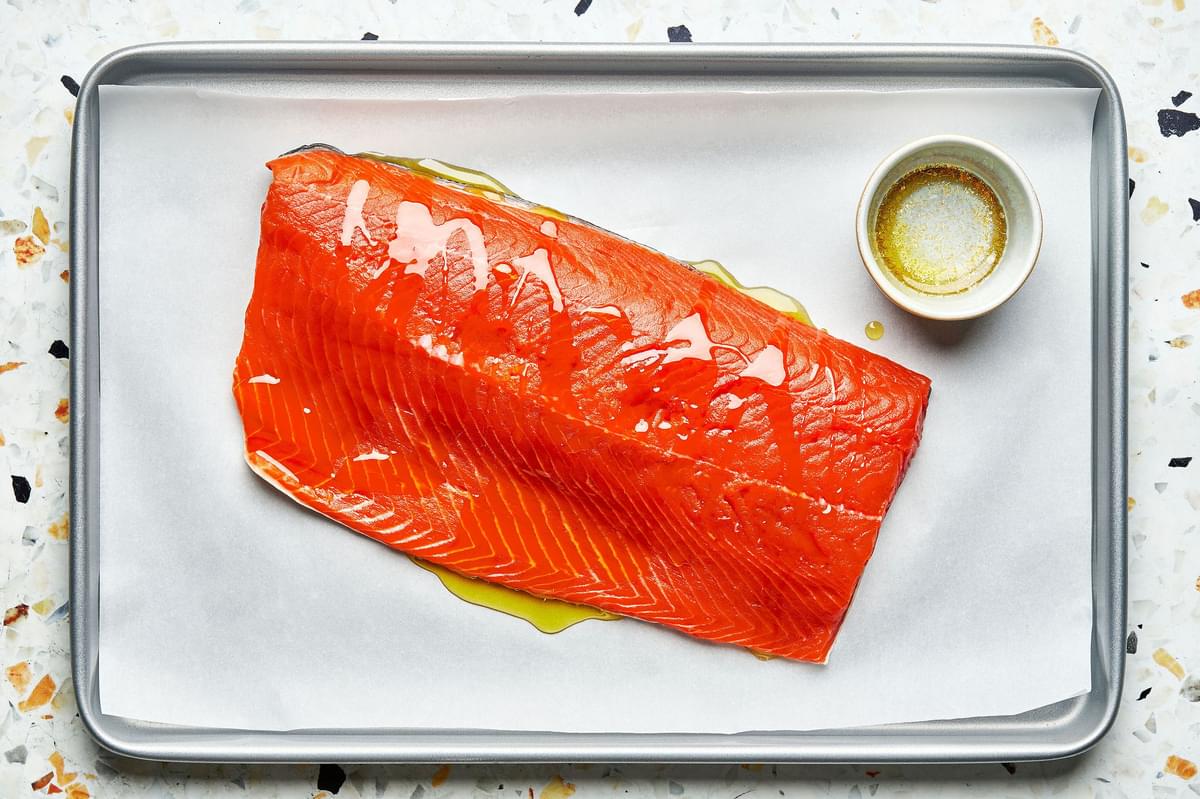 a salmon filet drizzled with olive oil on a parchment paper lined baking sheet next to a bowl of olive oil