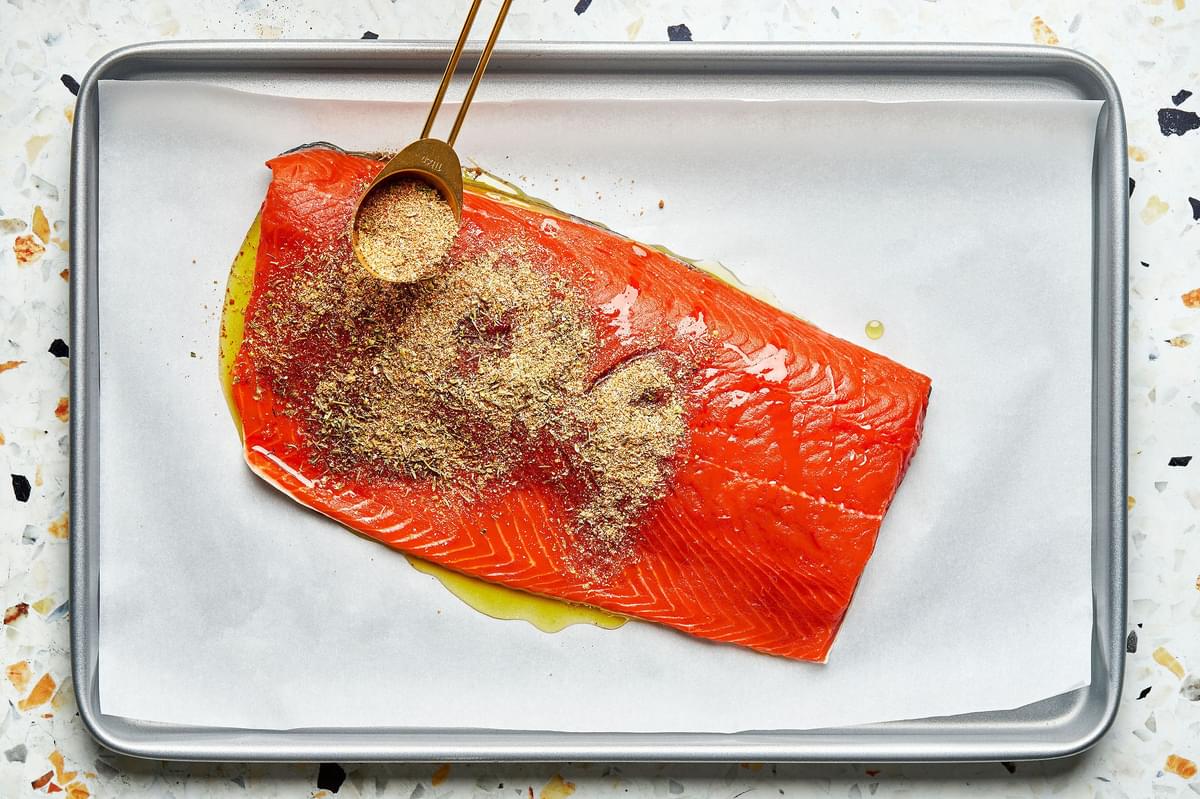 a salmon filet drizzled with olive oil on a parchment paper lined baking sheet being sprinkled with a spice rub
