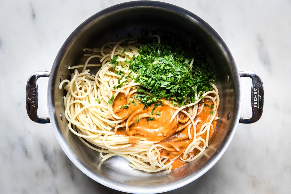 cooked bucatini noodles being tossed with spicy roasted red paper sauce and fresh parsley in a stock pot