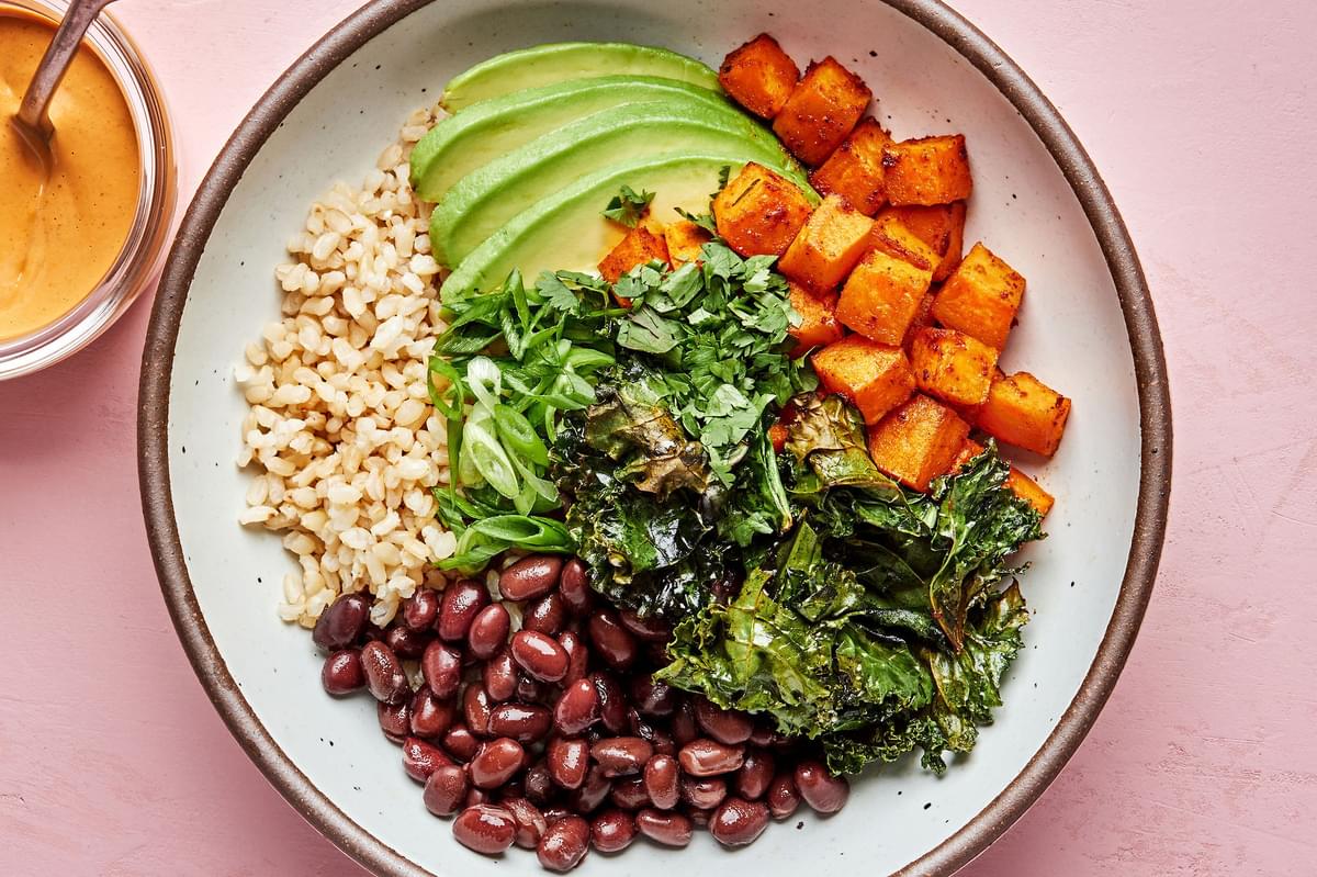 brown rice, black beans, avocado, roasted sweet potatoes and roasted kale in a bowl next to vegan cashew cheese sauce