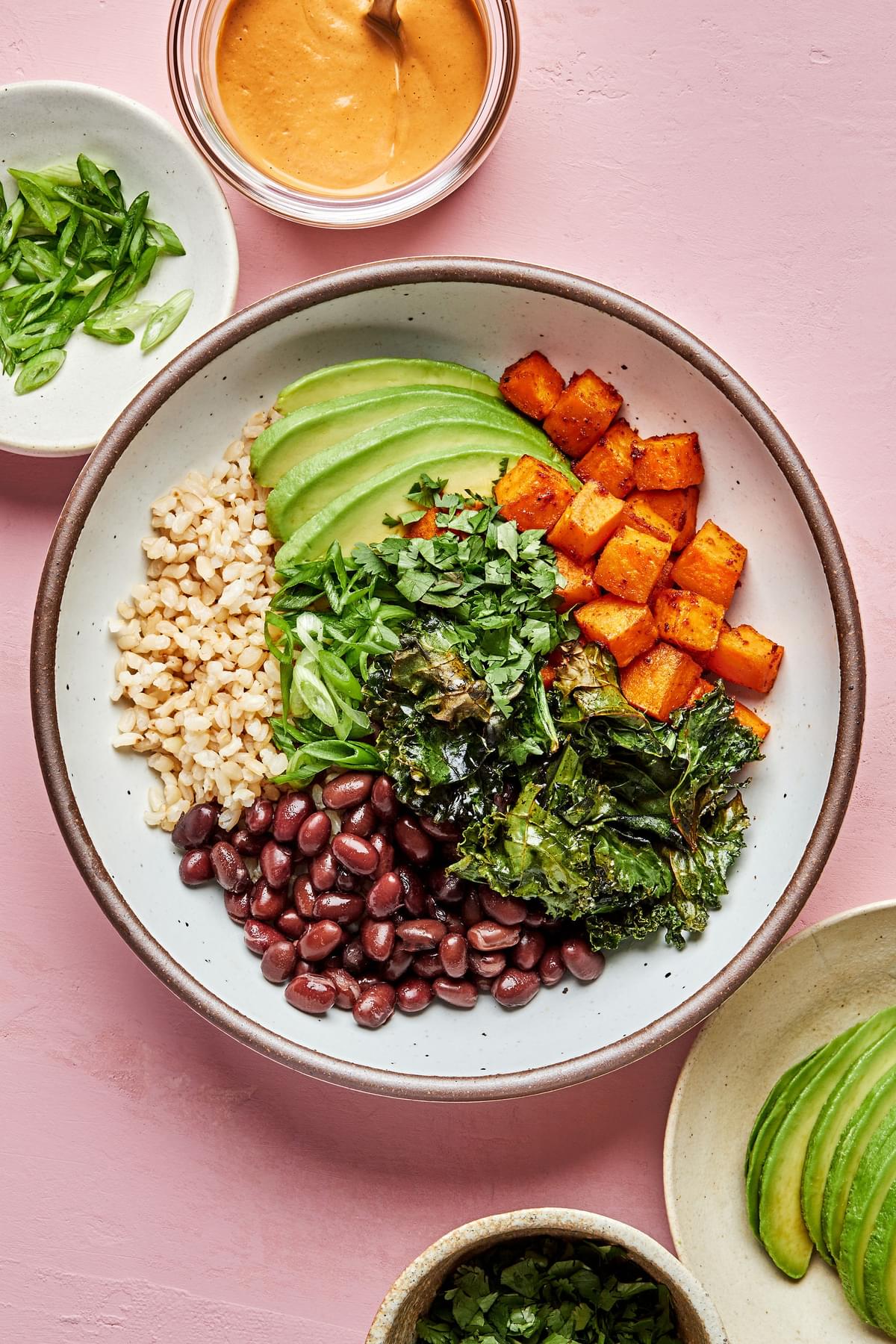 brown rice, black beans, avocado, roasted sweet potatoes and roasted kale in a bowl next to vegan cashew cheese sauce