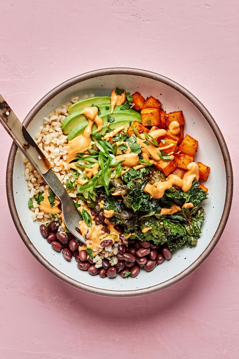 brown rice, black beans, avocado, roasted sweet potatoes and roasted kale in a bowl drizzled with vegan cashew cheese sauce