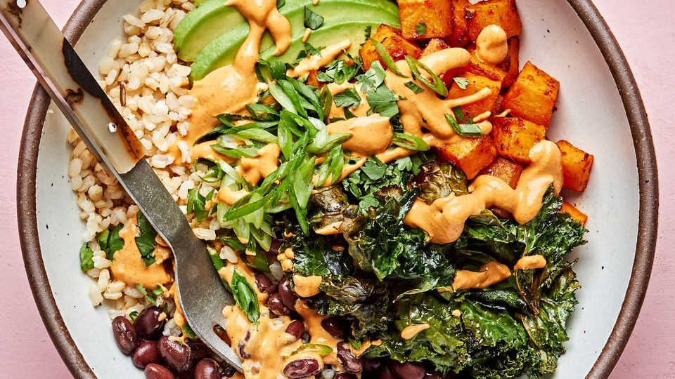 brown rice, black beans, avocado, roasted sweet potatoes and roasted kale in a bowl drizzled with vegan cashew cheese sauce
