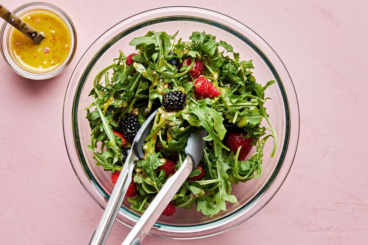 Arugula being tossed with summer berries and homemade balsamic vinaigrette beside a bowl of extra vinaigrette