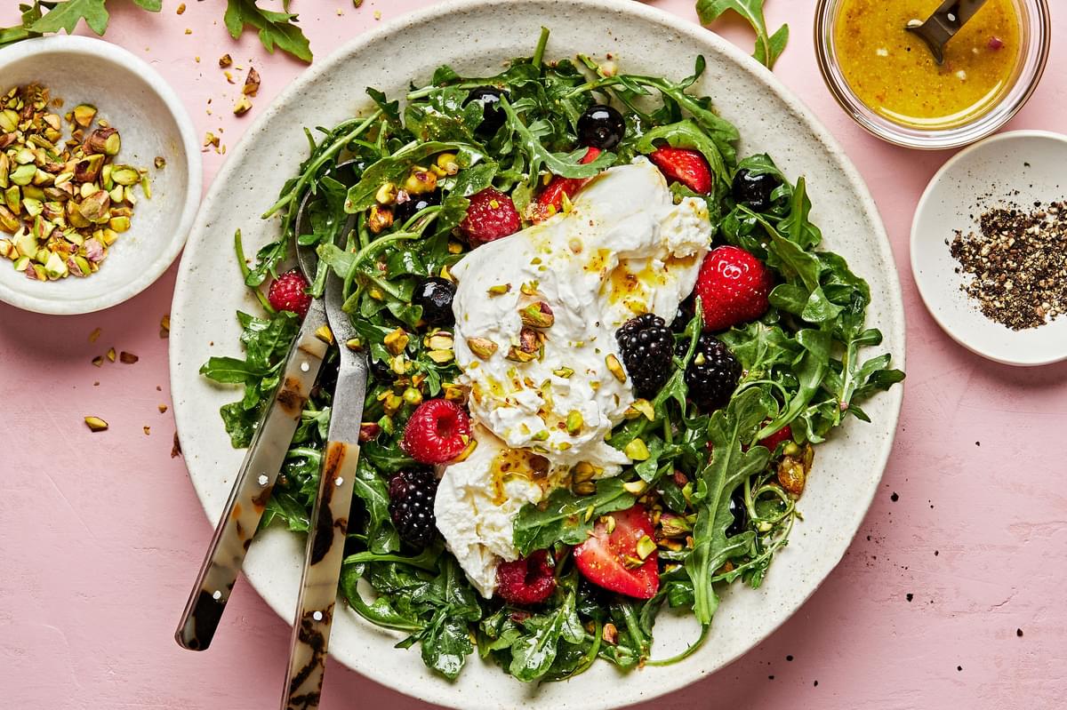 Burrata Salad with Summer Berries & Pistachios surrounded by bowls of pistachios, dressing and fresh cracked pepper
